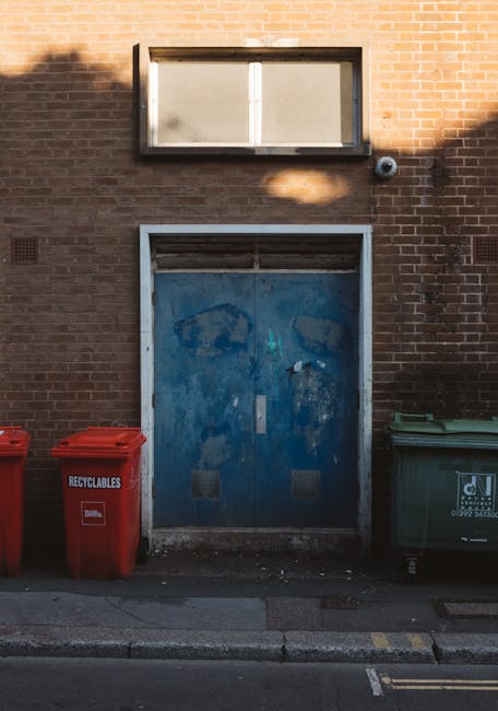 The image depicts a brick wall facade featuring a large, weathered blue metal door located near pavement level, with visible rust and paint chipping, indicating frequent use or age. Above the door, th